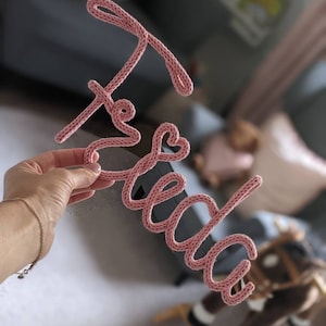 Könnte beinhalten: Ein pinkfarbenes, gestricktes Wort "Frida" wird in einer Hand gehalten. Das Wort besteht aus dickem Garn und ist in einer kursiven Schriftart geformt.