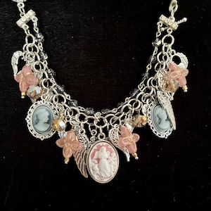 Puede incluir: Un collar de dijes plateado con varios colgantes. El collar presenta colgantes de camafeo, dijes de flores rosas, dijes de alas de &aacute;ngel y cuentas de cristal. El fondo es negro.
