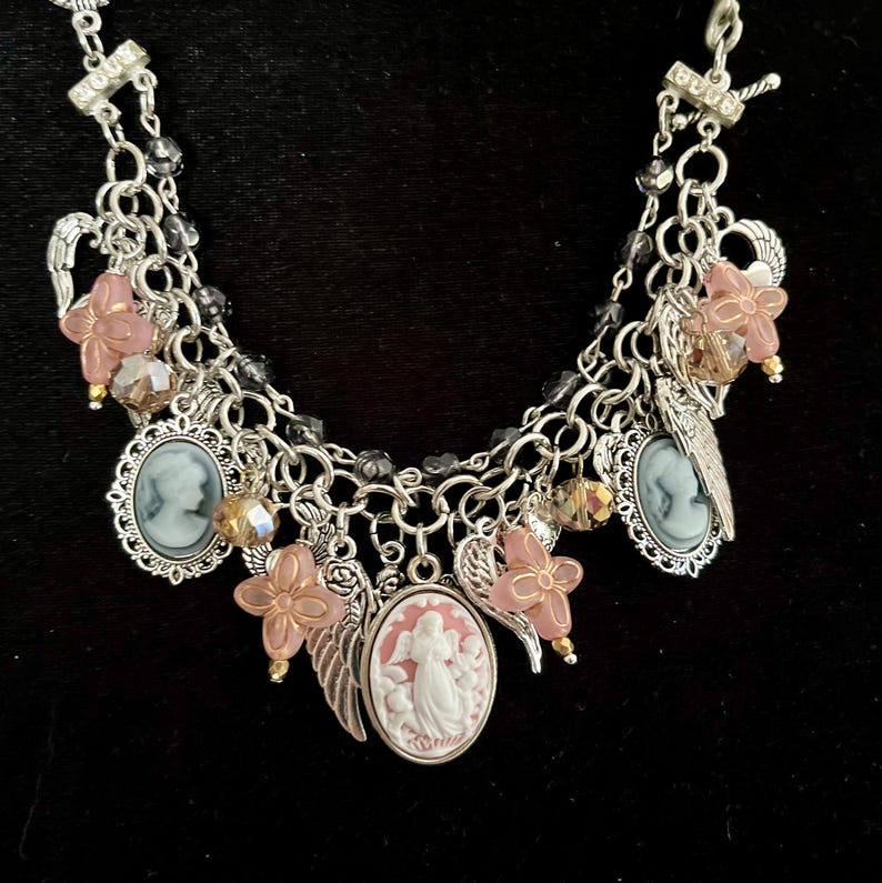 Puede incluir: Un collar de dijes plateado con varios colgantes. El collar presenta colgantes de camafeo, dijes de flores rosas, dijes de alas de &aacute;ngel y cuentas de cristal. El fondo es negro.