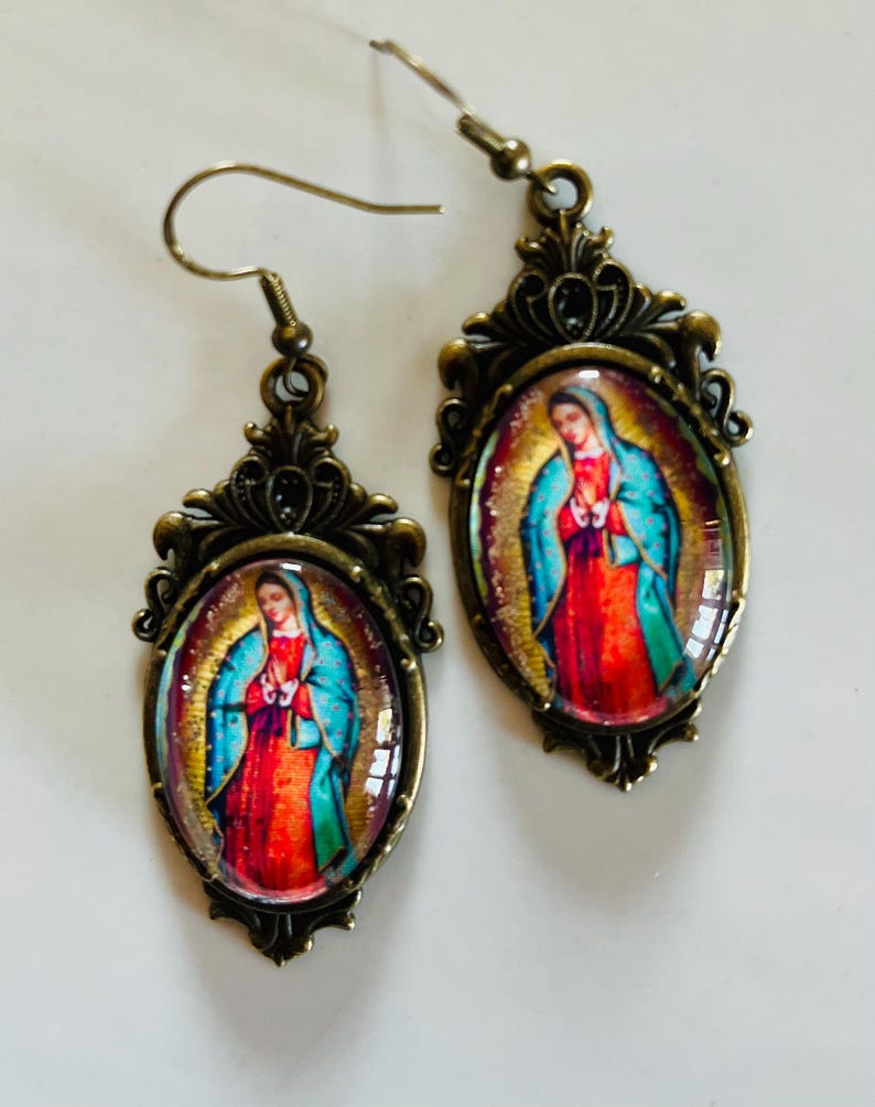 May include: A pair of bronze-coloured earrings with oval-shaped glass pendants featuring a colourful image of the Virgin Mary. The pendants have ornate metal frames.