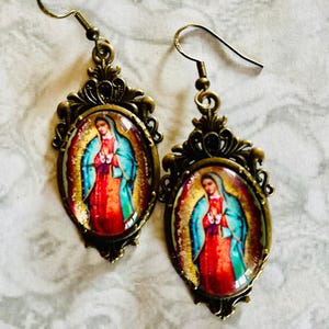 May include: A pair of gold-toned earrings with oval-shaped glass cabochons featuring an image of the Virgin Mary. The earrings have a detailed filigree design.