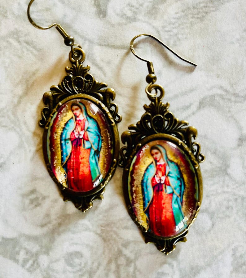 May include: A pair of gold-toned earrings with oval-shaped glass cabochons featuring an image of the Virgin Mary. The earrings have a detailed filigree design.