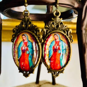 May include: A pair of gold-tone earrings with oval-shaped glass cabochons featuring a colourful image of the Virgin Mary. The earrings have a detailed ornate design.