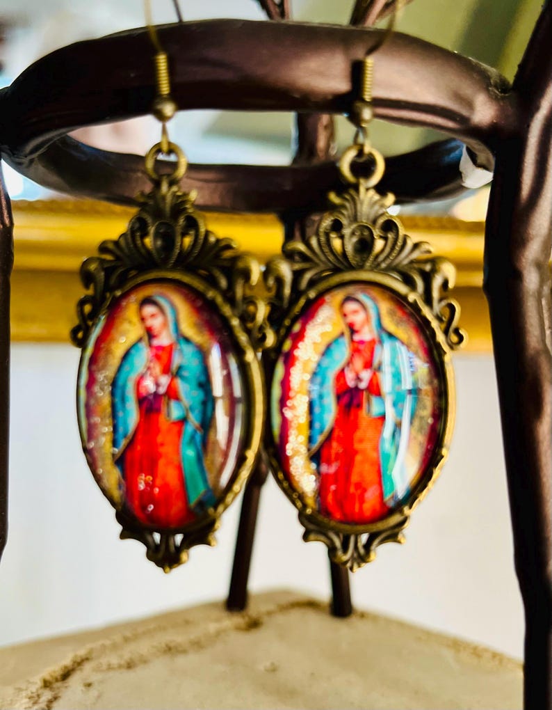 May include: A pair of gold-tone earrings with oval-shaped glass cabochons featuring a colourful image of the Virgin Mary. The earrings have a detailed ornate design.