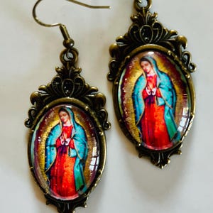 May include: A pair of gold-tone earrings featuring a cameo of Our Lady of Guadalupe. The cameo is set in an oval frame with a decorative filigree design.