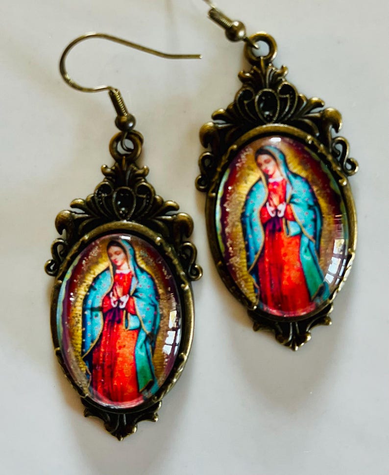 May include: A pair of gold-tone earrings featuring a cameo of Our Lady of Guadalupe. The cameo is set in an oval frame with a decorative filigree design.