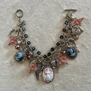 Puede incluir: Una pulsera de plata con m&uacute;ltiples colgantes y un cierre de palanca. La pulsera presenta varios colgantes, incluyendo colgantes de camafeo, acentos de flores rosas, alas de &aacute;ngel y colgantes en forma de coraz&oacute;n. La pulsera est&aacute; adornada con cuentas negras y detalles de cristal.