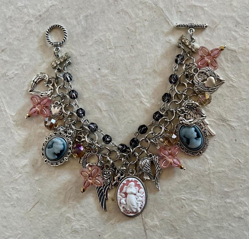 Puede incluir: Una pulsera de plata con m&uacute;ltiples colgantes y un cierre de palanca. La pulsera presenta varios colgantes, incluyendo colgantes de camafeo, acentos de flores rosas, alas de &aacute;ngel y colgantes en forma de coraz&oacute;n. La pulsera est&aacute; adornada con cuentas negras y detalles de cristal.