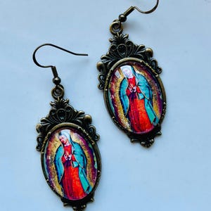 May include: A pair of gold-tone earrings with a vintage-style design. Each earring features a round, oval-shaped pendant with a colourful image of the Virgin Mary. The pendants are surrounded by ornate, filigree-like details.