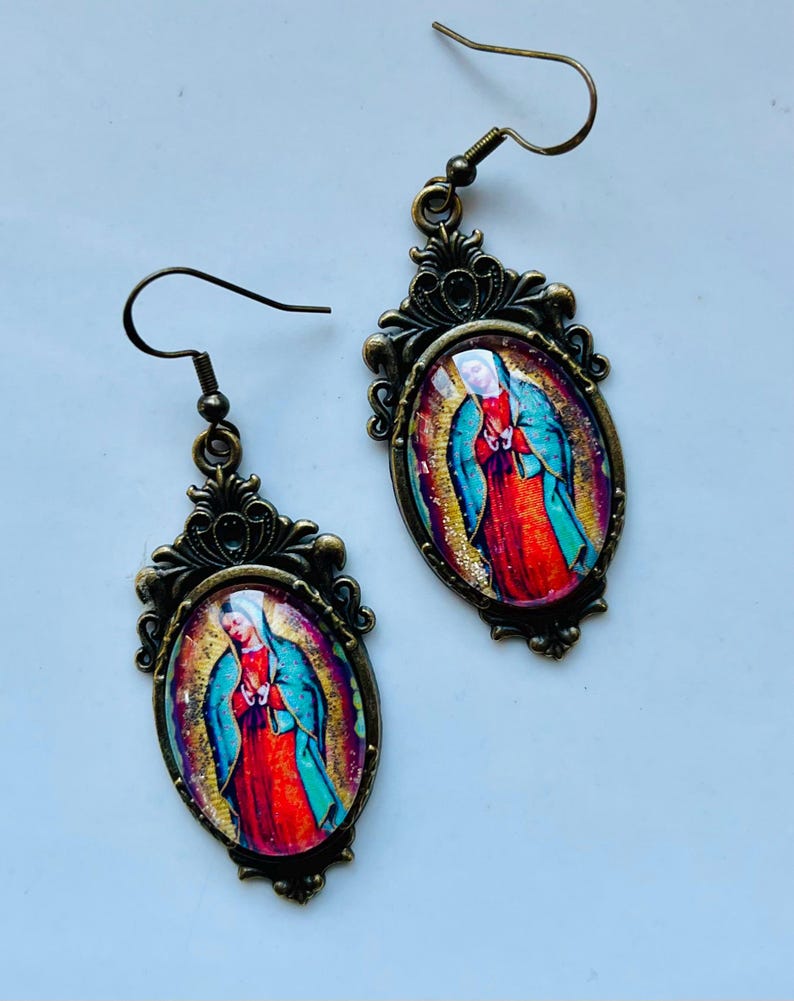 May include: A pair of gold-tone earrings with a vintage-style design. Each earring features a round, oval-shaped pendant with a colourful image of the Virgin Mary. The pendants are surrounded by ornate, filigree-like details.