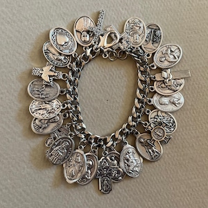 May include: A silver-toned charm bracelet featuring a thick curb chain adorned with numerous oval and cross-shaped metal charms. Each charm displays detailed embossed religious figures, symbols, and inscriptions, including St. Rosa and St. Benedict.