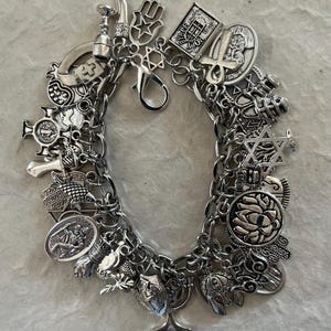 May include: A silver-toned charm bracelet featuring a variety of religious and spiritual symbols, including a Star of David, a hamsa hand, an ankh, a lotus flower, and a St. Christopher medal, all connected by a thick metal chain with a lobster clasp.