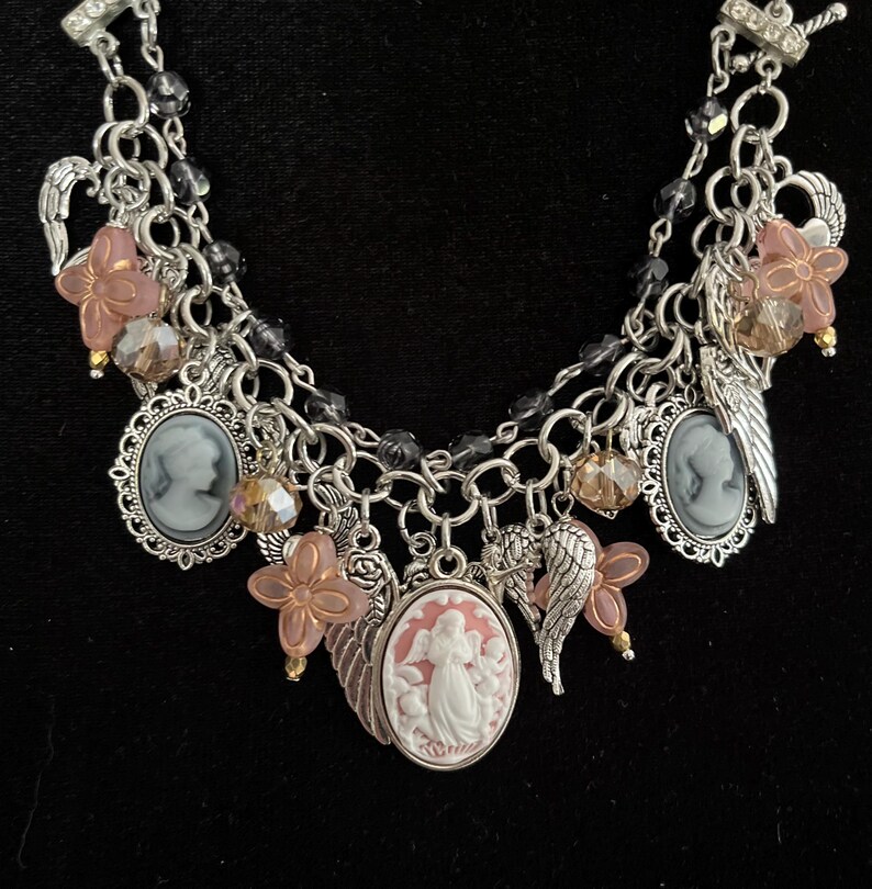 Puede incluir: Una pulsera de dijes plateada con m&uacute;ltiples cadenas y un cierre de palanca. La pulsera presenta colgantes de camafeo, dijes de alas de &aacute;ngel y detalles de flores rosas. Los dijes adicionales incluyen cuentas facetadas y peque&ntilde;as cuentas doradas. El fondo es negro.