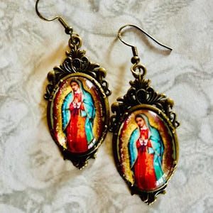 May include: A pair of gold-toned earrings with a detailed filigree design. Each earring features a round glass cabochon with a colourful image of the Virgin Mary.