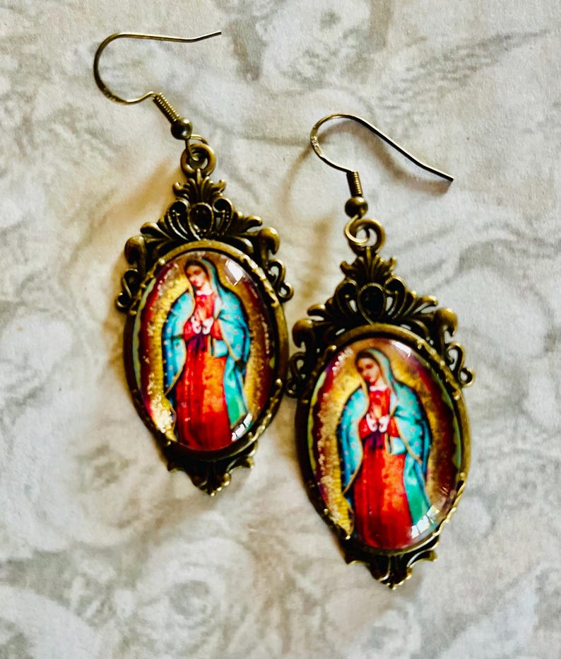 May include: A pair of gold-toned earrings with a detailed filigree design. Each earring features a round glass cabochon with a colourful image of the Virgin Mary.