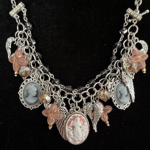 Puede incluir: Collar con colgantes plateados con m&uacute;ltiples colgantes. El collar presenta colgantes de camafeo, dijes de flores rosas y dijes de alas de &aacute;ngel. El collar tambi&eacute;n tiene detalles de cuentas transparentes y negras. Fondo negro.
