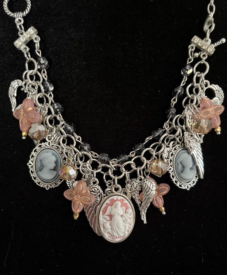 Puede incluir: Collar con colgantes plateados con m&uacute;ltiples colgantes. El collar presenta colgantes de camafeo, dijes de flores rosas y dijes de alas de &aacute;ngel. El collar tambi&eacute;n tiene detalles de cuentas transparentes y negras. Fondo negro.