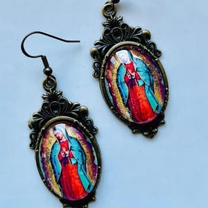 May include: A pair of gold-toned earrings with oval-shaped pendants featuring a colourful image of the Virgin Mary. The pendants have a detailed ornate design around the edges.