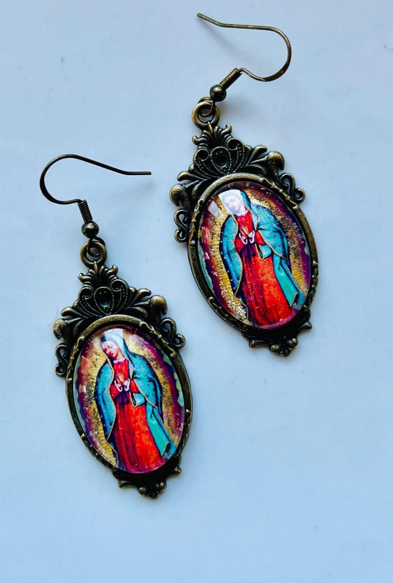 May include: A pair of gold-toned earrings with oval-shaped pendants featuring a colourful image of the Virgin Mary. The pendants have a detailed ornate design around the edges.