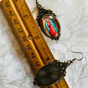 May include: A pair of bronze-toned earrings with oval-shaped glass insets. One earring features a colourful image of the Virgin Mary, while the other earring has a textured, sunburst design. The earrings are shown next to a ruler, measuring approximately 2.5 cm in length.