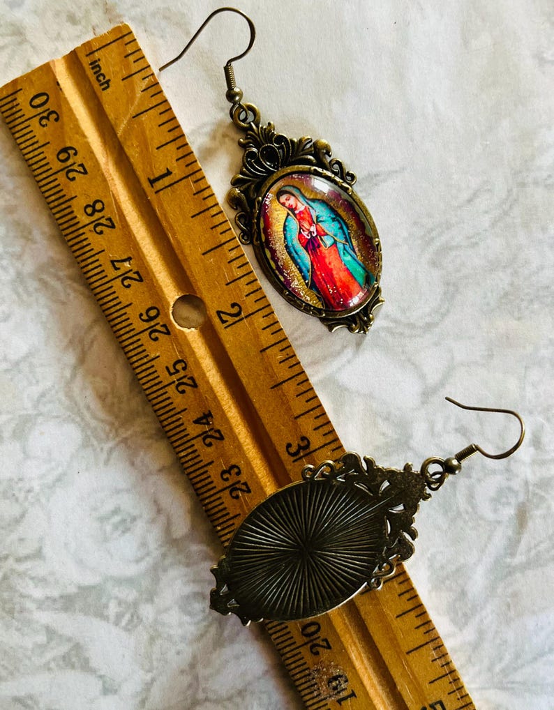 May include: A pair of bronze-toned earrings with oval-shaped glass insets. One earring features a colourful image of the Virgin Mary, while the other earring has a textured, sunburst design. The earrings are shown next to a ruler, measuring approximately 2.5 cm in length.