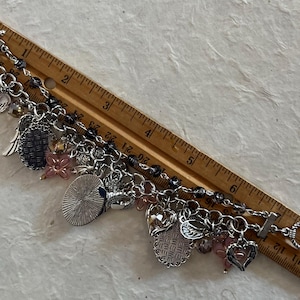 Puede incluir: Una pulsera de plata con varios dijes colgantes, incluyendo corazones, alas y flores. La pulsera mide aproximadamente 18 cm de largo y presenta un cierre de palanca. Cuentas rosas y transparentes se intercalan en el dise&ntilde;o.