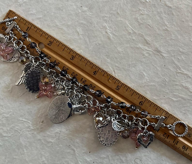 Puede incluir: Una pulsera de plata con varios dijes colgantes, incluyendo corazones, alas y flores. La pulsera mide aproximadamente 18 cm de largo y presenta un cierre de palanca. Cuentas rosas y transparentes se intercalan en el dise&ntilde;o.