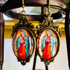 May include: A pair of gold-tone earrings featuring a religious image of the Virgin Mary in a blue and red robe. The earrings are oval-shaped and have an intricate design.