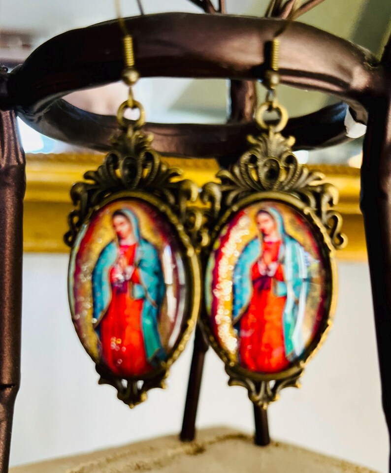 May include: A pair of gold-tone earrings featuring a religious image of the Virgin Mary in a blue and red robe. The earrings are oval-shaped and have an intricate design.