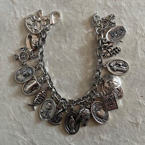 May include: Silver charm bracelet featuring various religious symbols, including crosses, saints, and other religious icons. The bracelet is made of a silver-colored metal and has a lobster clasp closure. The charms are detailed and vary in shape and size.