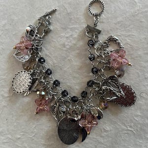 Puede incluir: Pulsera de abalorios plateada con cierre de palanca. La pulsera presenta m&uacute;ltiples cadenas y una variedad de abalorios, que incluyen cuentas de flores rosas, hojas plateadas y abalorios redondos y ovalados decorativos. Las cuentas negras se intercalan a lo largo de las cadenas.