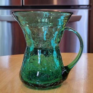 May include: A green crackle glass pitcher with a curved handle. The pitcher has a wide mouth and a rounded base. It sits on a light-colored wooden surface. The background includes a stainless steel refrigerator and wooden cabinets.