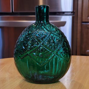 May include: A dark green glass vase with an intricate diamond and floral pattern. The vase has a rounded body and a narrow neck. The glass has a glossy finish, reflecting light. The vase is sitting on a light-colored wooden surface.