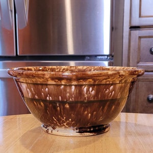 May include: A brown and white ceramic mixing bowl with a wide rim. The bowl has a speckled design and is sitting on a light-colored wooden surface. The background includes a stainless steel refrigerator and kitchen cabinets.