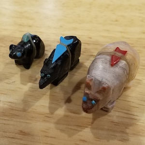 May include: Three small carved stone animal figurines. The figurines are a black bear, a black bear with a blue turquoise saddle, and a tan bear with a red turquoise saddle. The bears are all carved in a realistic style and have small turquoise eyes.