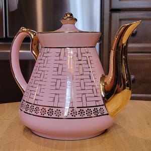 May include: A pink ceramic teapot with gold accents. The teapot features a woven pattern and a floral design around the base. The handle and spout are gold-colored. The teapot is sitting on a wooden surface.