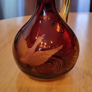 May include: A red glass decanter with a gold stopper and a carved design of a bird in flight. The decanter has a small handle and a round body.