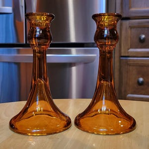 May include: Two amber-colored glass candle holders. Each has a flared base, a slender stem, and a cup-shaped top. The candle holders are placed on a light-colored wooden surface.