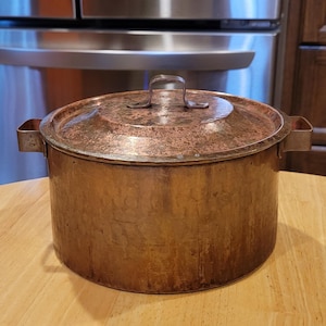 May include: A copper pot with a lid and two handles. The pot is on a wooden table.