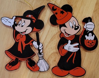 May include: Two Halloween-themed wooden ornaments featuring Mickey and Minnie Mouse. Mickey is dressed as a wizard with a red hat and a black robe, holding a pumpkin-shaped bag. Minnie is dressed as a witch with a black hat and a black dress, holding a broom.