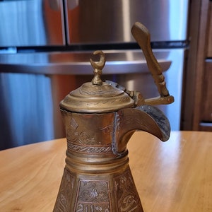 May include: Antique brass coffee pot with intricate engraved designs and a hinged lid. The pot features a long spout and a wooden handle. The pot is sitting on a wooden table.