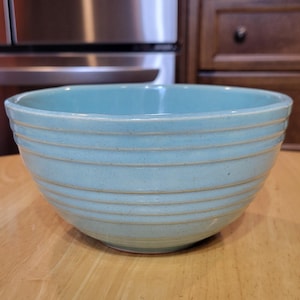 May include: A light blue ceramic mixing bowl with horizontal ridges. The bowl is set on a light brown wooden surface. The background includes a stainless steel refrigerator and wooden cabinets.