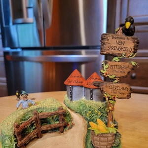 May include: A decorative figurine featuring a rural scene with a path, fence, and signposts. The signposts direct to New Springfield, Pittsburgh, and Cleveland. A basket of corn and a small crow are also present. The figurine has the text "Hummel Gift Shop".