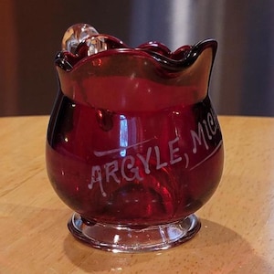 May include: A small, red glass creamer with a handle and a white inscription that reads "ARGYLE, MI".