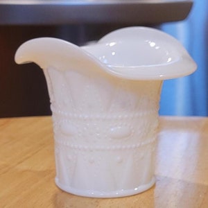 May include: A white milk glass creamer with a scalloped rim and embossed design. The creamer is sitting on a wooden table.