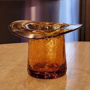 May include: An amber-colored glass top hat-shaped container. The glass has a textured, hammered appearance. The container sits on a light-colored wooden surface. The background includes a stainless steel refrigerator and wooden cabinets.