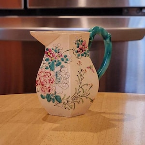 May include: A white ceramic pitcher with a green handle and floral design. The pitcher has pink, green, and white flowers painted on it.