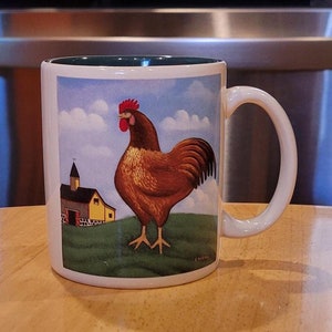 May include: White ceramic coffee mug with a green rim. The mug features a colorful illustration of a rooster standing in a field with a small yellow building in the background.