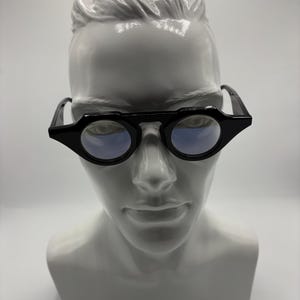 May include: Black eyeglasses with circular lenses and a thick, angular frame bridge. The glasses feature a distinct geometric design with pointed outer edges, resting on a white mannequin head. The lenses have a subtle blue tint and a reflective finish.