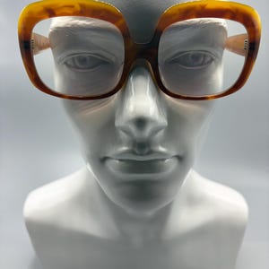 May include: Oversized square-framed spectacles with a tortoiseshell design. The glasses are presented on a white bust, highlighting their striking shape and retro style. The frames feature a warm, brown and amber colour.